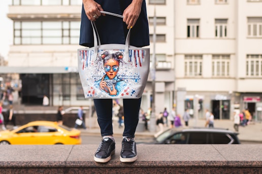 A person holding a tote bag with an artistic design featuring a girl with blue sunglasses and hair buns. The bag is white with colorful accents, including red and blue paint drips. The background shows a busy urban street scene with blurred pedestrians and a moving yellow car.