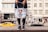 A person holding a tote bag with an artistic design featuring a girl with blue sunglasses and hair buns. The bag is white with colorful accents, including red and blue paint drips. The background shows a busy urban street scene with blurred pedestrians and a moving yellow car.