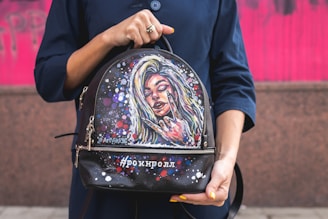 A person holds a backpack with a colorful painted design featuring a woman's face in a vibrant, artistic style. The background is a bright pink wall, and the person is wearing a dark blue outfit. The backpack features various colorful accents and graffiti-style lettering.