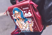 A leather bag with a pop art illustration of a surprised woman with blue hair and red glasses holding a takeaway coffee cup. The bag is red with white dots and has text written in Cyrillic script. The person carrying it is wearing a black leather jacket.