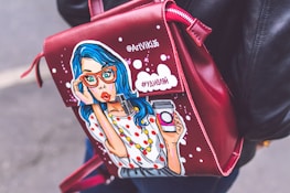 A leather bag with a pop art illustration of a surprised woman with blue hair and red glasses holding a takeaway coffee cup. The bag is red with white dots and has text written in Cyrillic script. The person carrying it is wearing a black leather jacket.