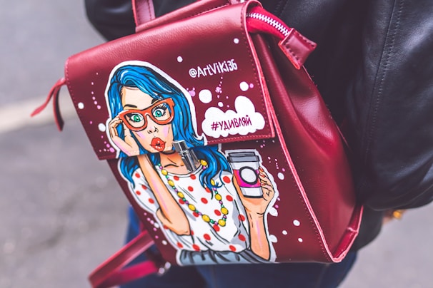 A leather bag with a pop art illustration of a surprised woman with blue hair and red glasses holding a takeaway coffee cup. The bag is red with white dots and has text written in Cyrillic script. The person carrying it is wearing a black leather jacket.