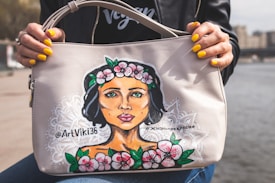 A person holds a handbag decorated with an artistic portrait of a woman with flowers in her hair. The woman's hair is dark, and the flowers are pink with green leaves. The handbag is light-colored, and the person holding it has painted yellow nails.