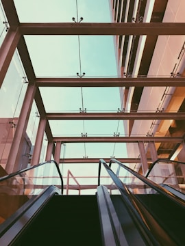 A modern architectural setting featuring a pair of escalators leading upward towards a glass ceiling. The structure of the ceiling includes metal beams and clear glass panels that reveal a view of the sky above. The scene captures geometric lines and reflections, creating a sense of symmetry and openness.