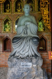 A stone statue of a monk holding prayer beads is placed against a brick wall with gold-framed niches. Each niche contains small Buddha figures, adding a spiritual ambiance. The wall also features intricate golden carvings.