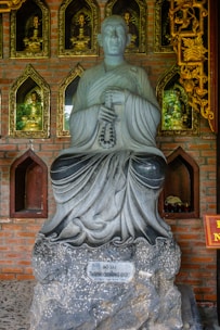 A stone statue of a monk holding prayer beads is placed against a brick wall with gold-framed niches. Each niche contains small Buddha figures, adding a spiritual ambiance. The wall also features intricate golden carvings.