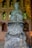 A stone statue of a monk holding prayer beads is placed against a brick wall with gold-framed niches. Each niche contains small Buddha figures, adding a spiritual ambiance. The wall also features intricate golden carvings.