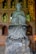 A stone statue of a monk holding prayer beads is placed against a brick wall with gold-framed niches. Each niche contains small Buddha figures, adding a spiritual ambiance. The wall also features intricate golden carvings.