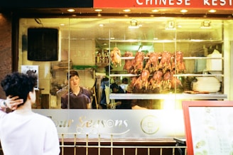 A person stands outside a Chinese restaurant, looking through a glass window display. Inside, several roast ducks are hung, with a restaurant worker visible in the background. The bright lighting highlights the cooked meats and the interior of the restaurant.