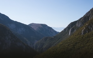 The landscape depicts a mountain range with rugged terrain. The mountains are covered with a mix of dense green forests and rocky outcrops. The sky above is clear, adding a sense of serenity and vastness to the scene. The sunlight creates gentle contrasts on the slopes, highlighting the natural textures and contours of the mountains.