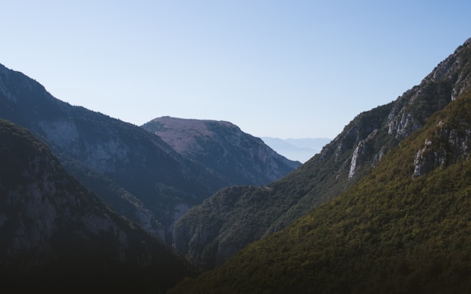The landscape depicts a mountain range with rugged terrain. The mountains are covered with a mix of dense green forests and rocky outcrops. The sky above is clear, adding a sense of serenity and vastness to the scene. The sunlight creates gentle contrasts on the slopes, highlighting the natural textures and contours of the mountains.