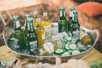 A metal oval tray is filled with ice, bottles of beer, and slices of cucumber. The beer bottles are both green and clear with labels, some of which are partially covered by frost. The setting appears to be outdoors with a blurred background featuring earthy tones.