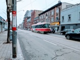 A city street features a vintage red and white streetcar traveling down tracks. The surroundings include a mix of brick buildings and modern architecture on either side of the street. Several cars are parked along the sidewalk, and a few pedestrians are visible. A bus stop sign with route information is situated in the foreground, next to a tree.