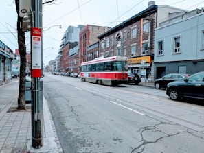 A city street features a vintage red and white streetcar traveling down tracks. The surroundings include a mix of brick buildings and modern architecture on either side of the street. Several cars are parked along the sidewalk, and a few pedestrians are visible. A bus stop sign with route information is situated in the foreground, next to a tree.