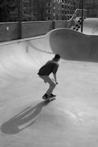 A skateboarder is captured in mid-motion at a skate park, maneuvering over a concrete ramp. The scene is in black and white, highlighting the play of shadows on the smooth surface. In the background, brick walls and trees are visible, framed by residential buildings.