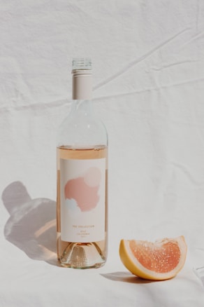 A bottle of ros&eacute; wine with a minimalist label stands upright on a soft, textured fabric backdrop. Beside it lies a slice of pink grapefruit, placed to the right, casting a gentle shadow. The overall composition is lightly bathed in natural sunlight, giving the scene an airy, fresh feel.