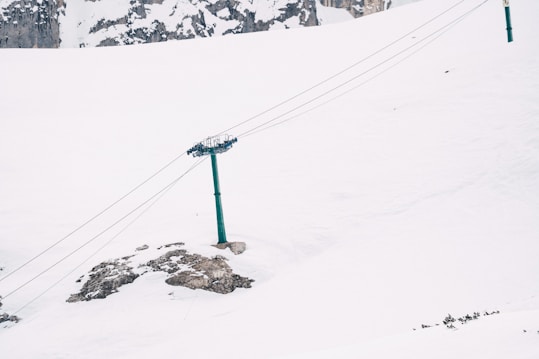 A snow-covered landscape featuring a ski lift structure with cables extending across the scene. The lift pole stands on a hill with snowy mounds and some exposed rock. In the background, the terrain slopes gently, and there are distant rocky cliffs.