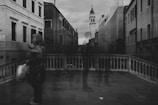 A black and white photograph of an urban street scene with ghostly figures and old buildings. The architecture features ornate facades and a bell tower in the background. One person with a blurred effect is carrying a bag, walking across a stone bridge with a balustrade.