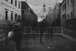 A black and white photograph of an urban street scene with ghostly figures and old buildings. The architecture features ornate facades and a bell tower in the background. One person with a blurred effect is carrying a bag, walking across a stone bridge with a balustrade.