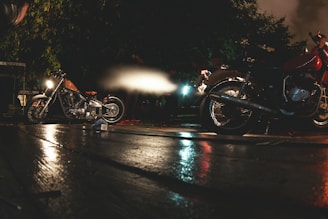 Two motorcycles are parked on a wet surface reflecting light. The scene is set at night, surrounded by dark trees and dim lighting, creating a moody, atmospheric setting.