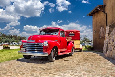 A classic, vintage red pickup truck with chrome accents is parked on a cobblestone path. It is towing a red and yellow trailer. The sky is clear with a few scattered clouds, and there is a rustic building nearby. The surrounding area includes green grass and some trees.