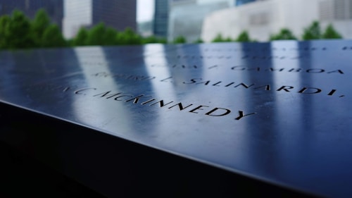 An engraved memorial plaque with names inscribed, placed in an outdoor setting surrounded by green trees and blurred city buildings in the background.