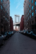 A view down a cobblestone street flanked by brick buildings with parked cars on both sides. In the background, the Manhattan Bridge is visible framed between the buildings, with the skyline faintly visible beyond.