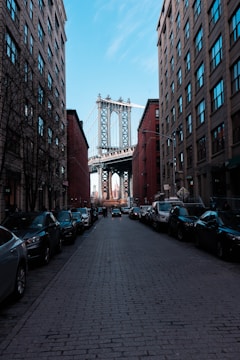 A view down a cobblestone street flanked by brick buildings with parked cars on both sides. In the background, the Manhattan Bridge is visible framed between the buildings, with the skyline faintly visible beyond.