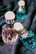 Several elegant perfume bottles with textured surfaces and circular caps are displayed on a soft fabric background. The bottles vary in color, including pink and teal, with prominent branding on the cap's band.