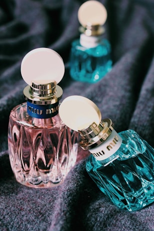 Several elegant perfume bottles with textured surfaces and circular caps are displayed on a soft fabric background. The bottles vary in color, including pink and teal, with prominent branding on the cap's band.
