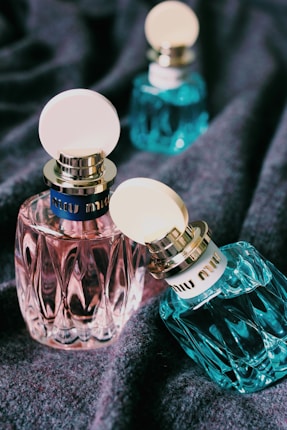 Several elegant perfume bottles with textured surfaces and circular caps are displayed on a soft fabric background. The bottles vary in color, including pink and teal, with prominent branding on the cap's band.