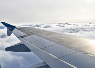 An airplane wing is visible stretching across the image, with fluffy clouds below and a bright sky above. The wing features various panels and a sleek design.