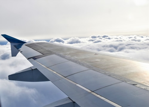 An airplane wing is visible stretching across the image, with fluffy clouds below and a bright sky above. The wing features various panels and a sleek design.
