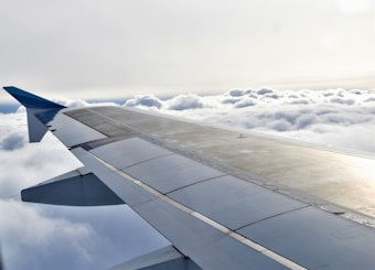 An airplane wing is visible stretching across the image, with fluffy clouds below and a bright sky above. The wing features various panels and a sleek design.