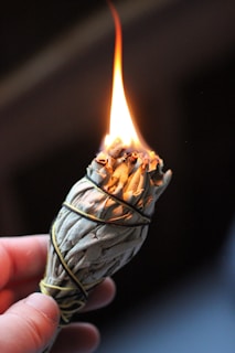 A person holds a smoldering bundle of sage wrapped in string, with a bright flame at the top. The focus is on the burning sage, with the background being dark and out of focus.