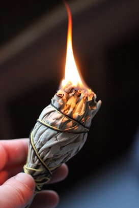 A person holds a smoldering bundle of sage wrapped in string, with a bright flame at the top. The focus is on the burning sage, with the background being dark and out of focus.