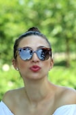 A person wearing sunglasses with reflective lenses, capturing a serene outdoor setting with greenery in the background. The individual has pulled back hair and is dressed in a white off-the-shoulder top, while displaying a playful expression with puckered lips.