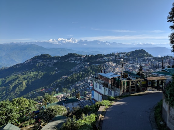 A scenic view of a hilly town with a winding road in the foreground, surrounded by lush greenery and scattered houses. In the background, a range of majestic snow-capped mountains can be seen under a clear blue sky, adding depth to the landscape.
