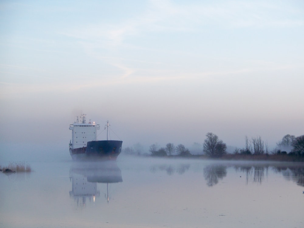 blue and white cargo ship sailing on calm glassy canal with fogs