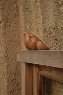 A wooden bird sculpture is placed on a rough wooden ledge beside a textured wall. The sculpture is elegantly crafted with smooth curves and a polished finish, contrasting with the raw, earthy tones of the background.