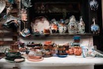 A wide array of handcrafted ceramic items, including mugs, cups, and vases, are displayed on a table. The ceramics are painted with vibrant, mixed colors including blues, oranges, and greens, and feature intricate patterns and designs. There are artistic depictions of animals like owls and elephants, alongside abstract shapes and forms. The display also includes hanging decorative items like a fish and other ornate figures.