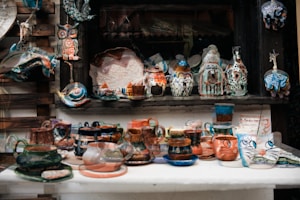 A wide array of handcrafted ceramic items, including mugs, cups, and vases, are displayed on a table. The ceramics are painted with vibrant, mixed colors including blues, oranges, and greens, and feature intricate patterns and designs. There are artistic depictions of animals like owls and elephants, alongside abstract shapes and forms. The display also includes hanging decorative items like a fish and other ornate figures.