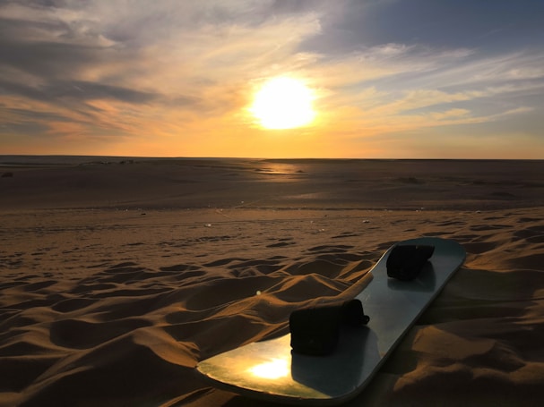 A serene desert landscape at sunset, with the sun low on the horizon, casting a warm, golden glow over the sand. In the foreground, a sandboard rests on the dunes, suggesting a sense of adventure and tranquility.