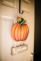 A decorative wooden pumpkin with textured edges and painted autumn leaves is hanging on a beige door. Below the pumpkin, a wooden sign with the word 'Welcome' written in white cursive is attached.