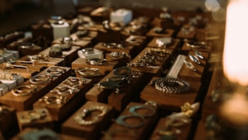 A collection of various pieces of jewelry, including rings, bracelets, and necklaces, displayed on wooden blocks. The arrangement is illuminated with warm lighting, creating a cozy and inviting atmosphere.