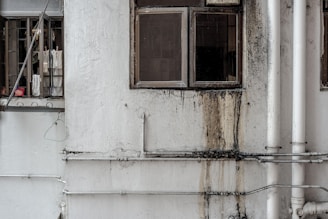 A worn-out facade of a building featuring two windows. One window is partially open with laundry hanging, including shirts and a pink basin visible inside. The wall is stained with grime and water damage near the other window, with multiple white pipes running vertically and horizontally across the surface.