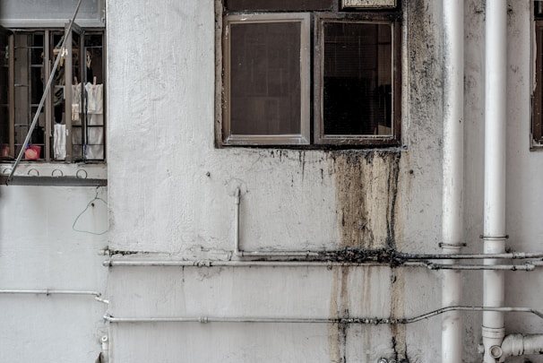 A worn-out facade of a building featuring two windows. One window is partially open with laundry hanging, including shirts and a pink basin visible inside. The wall is stained with grime and water damage near the other window, with multiple white pipes running vertically and horizontally across the surface.