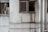 A worn-out facade of a building featuring two windows. One window is partially open with laundry hanging, including shirts and a pink basin visible inside. The wall is stained with grime and water damage near the other window, with multiple white pipes running vertically and horizontally across the surface.