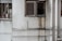 A worn-out facade of a building featuring two windows. One window is partially open with laundry hanging, including shirts and a pink basin visible inside. The wall is stained with grime and water damage near the other window, with multiple white pipes running vertically and horizontally across the surface.