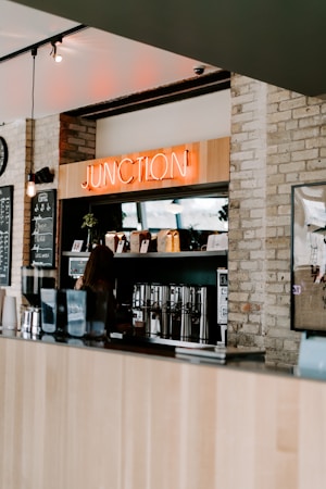 A cozy coffee shop interior with a wooden counter and a neon sign reading 'JUNCTION' mounted above a coffee station. Various coffee-making equipment and bags of coffee beans are neatly arranged on shelves. The walls are made of exposed brick, adding a rustic touch.